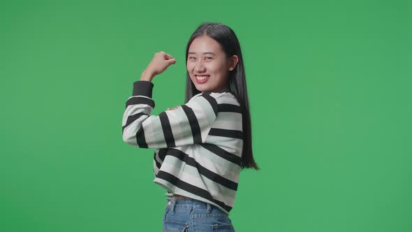 Side View Of A Smiling Asian Woman Flex Muscle While Standing In Front Of Green Screen Background