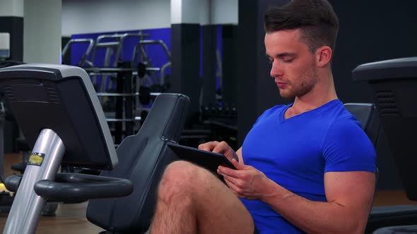 A Young Fit Man Trains on a Recumbent Bike in a Gym and Works on a Tablet - Closeup