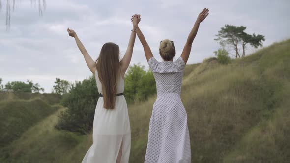 Back View of Two Slim Gorgeous Caucasian Women Standing Outdoors on Windy Day in Light White Dresses
