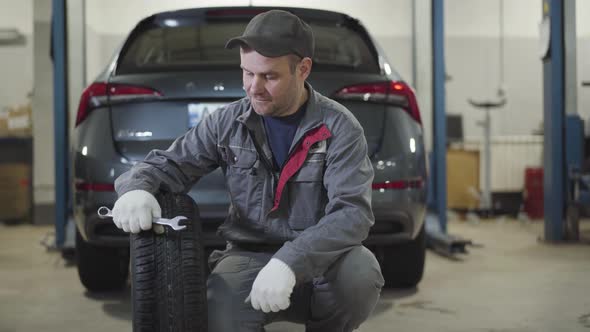 Portrait of Positive Caucasian Man Sitting with Wheel and Adjustable Wrench in Front of Car and