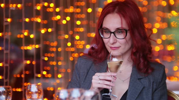 A Woman with Red Hair Sitting By Table in Restaurant and Drinking Champagne