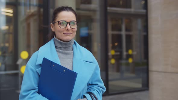 Smiling Businesswoman Wearing Blue Coat Holding Clipboard and Walking Outdoors