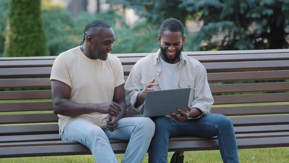 Two African American Male Friends or Colleagues Sitting on Bench in City Park Using Laptop