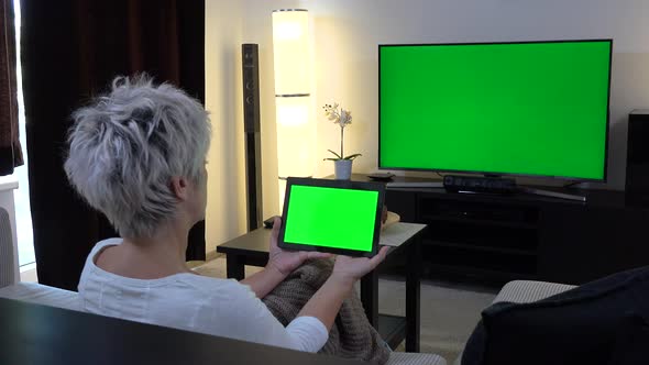 A Middle-aged Woman Sits, Watches TV with a Green Screen and Looks at a Tablet with a Green Screen