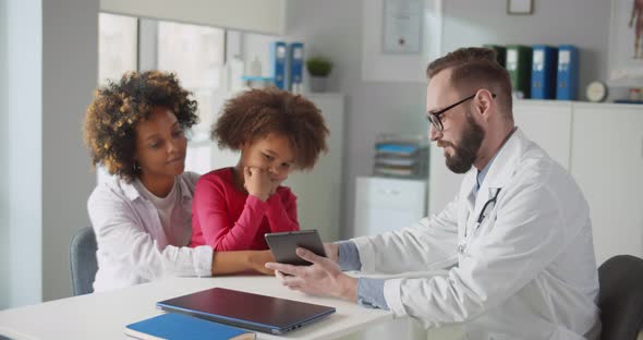 Young Male Doctor Showing Medical Test Results on Tablet to Young Woman with Little Daughter