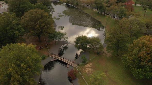 An aerial drone shot over a park in the day with a pond, with an arched footbridge. It's a beautiful