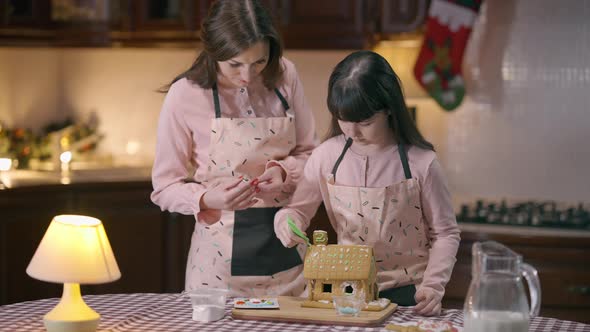 Portrait of Little Cute Girl Decorating Ginger House with Sugar Icing Standing in Kitchen with Woman