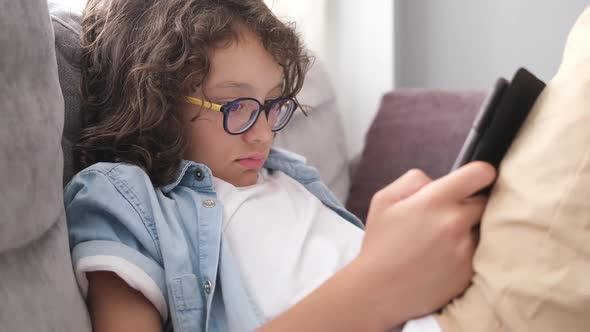 Little Cute Boy with Eyeglasses Sitting on Sofa and Playing Game on Digital Tablet Closeup Portrait