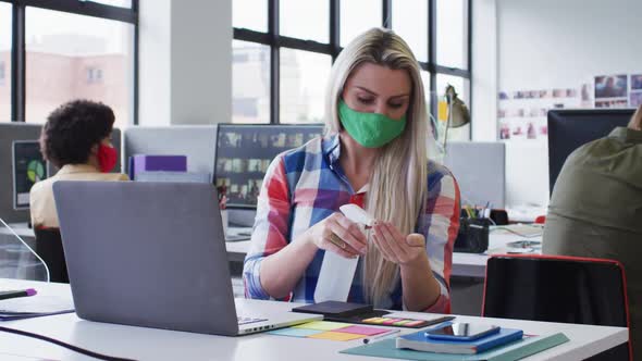 Caucasian businesswoman wearing face mask disinfecting hands using a laptop in modern office