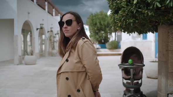 Portrait  of woman with sunglasses  in a beige trench coat against baby carriage