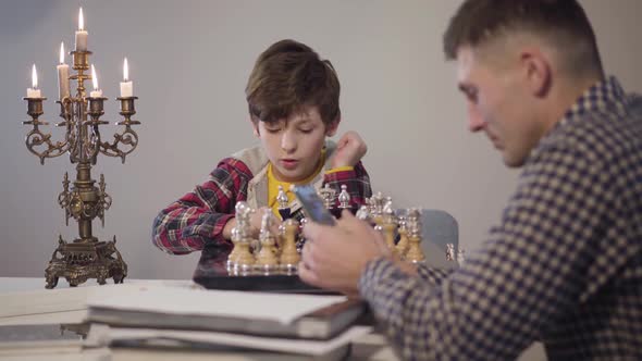 Portrait of Cute Smart Caucasian Boy Placing Chess on Board and Talking To Father Sitting at the