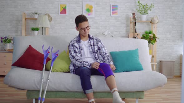 Young Asian Man Sitting on the Couch Puts a Tightening Elastic Bandage on a Sick Knee