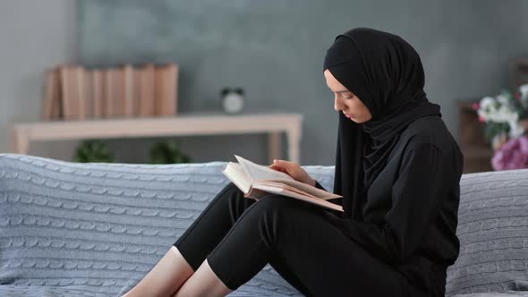 Focused Arabic Woman in Traditional Black Hijab Reading Book Relaxing on Couch