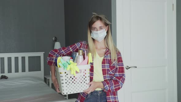 Woman with Mask on Face and Basket Full of Sponges and Household Chemicals Look at Camera