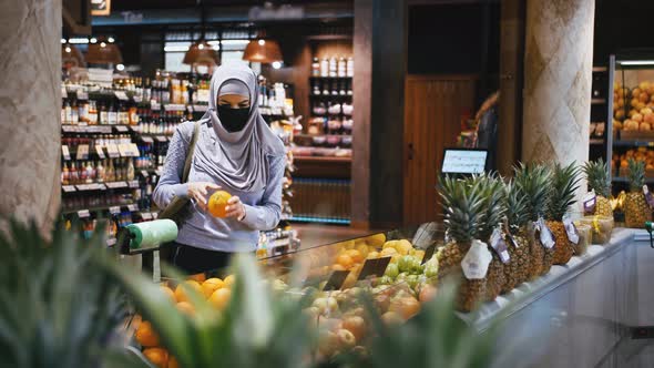 Young Beautiful Muslim Woman in Hijab and Protective Mask Choosing Fruits in the Supermarket and Put