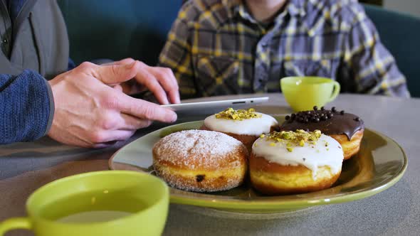 Two People Pointing At Tablet Computer Screen At Restaurant Table With Donuts