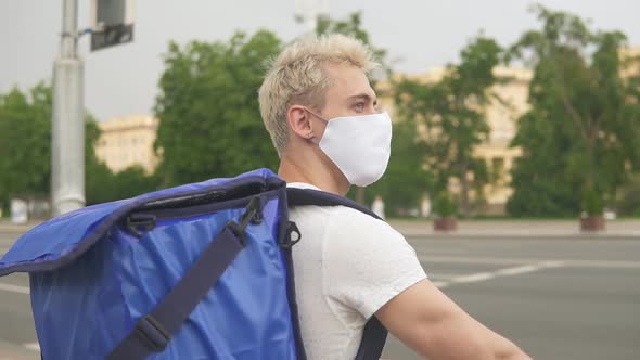 Male courier in medical mask delivering food with backpack, riding a bicycle.