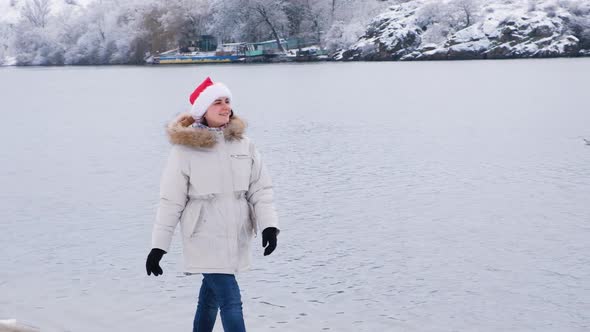 A Woman in a Santa Claus Hat Walks Along the Bank Along the River