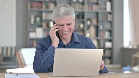 Middle Aged Man Talking on Smartphone While Working on Laptop in Office