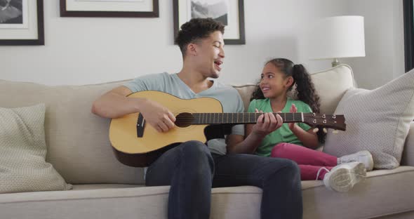 Happy biracial father and daughter sitting on sofa playing guitar