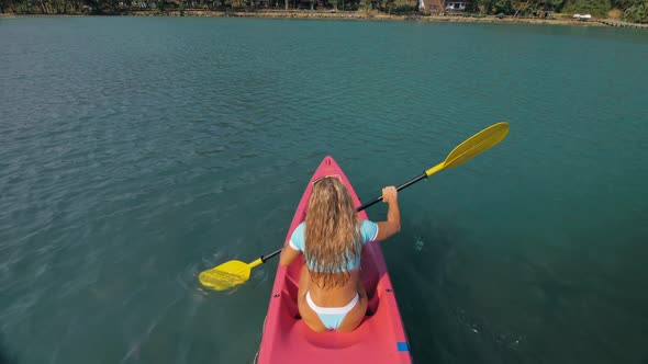 Seductive Blonde Woman in Blue Swimsuit Sails on Pink Plastic Kayak Along Azure Sea at Exotic Resort