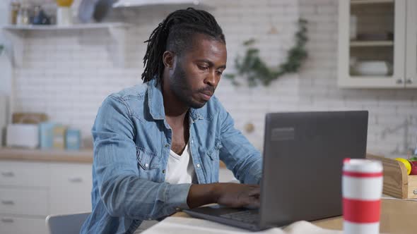 Concentrated African American Male Freelancer Typing on Laptop Keyboard Sitting in Kitchen at Home