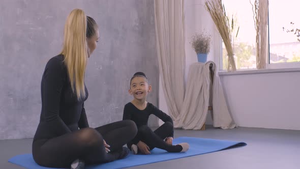 Beautiful Young Gymnastics Teacher and Little Asian Girl Sitting on Yoga Mat in Studio and Talking