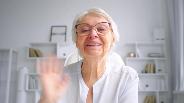 Old woman with grey hairstyle in glasses waves hands talking to close friend at online video