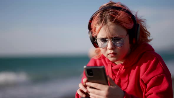 Preteen Girl is Viewing Learning Video in Smartphone Listening to Sound By Headphones on Sea Beach