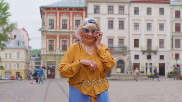 Senior Old Tourist Woman in Stylish Clothes Talking on Mobile Phone While Walking on City Street