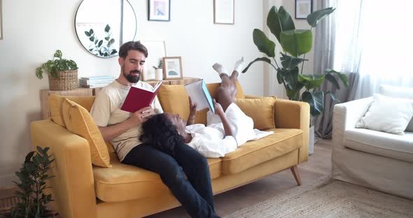 Multiethnic Couple Reading a Book Lying on the Sofa at Home in an Apartment