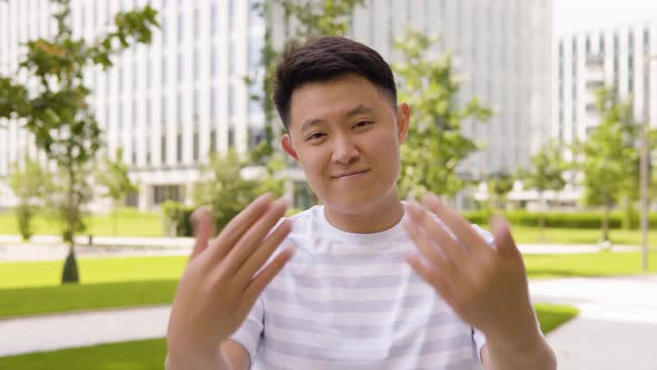 A Young Asian Man Waves at the Camera in a Gesture of Invitation with a Smile in an Urban Area