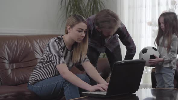 Cute Young Woman Sitting on the Leather Sofa and Bearded Man Standing Near Her Working with Laptop