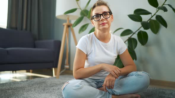 Woman with Glasses Looking at the Camera Sitting on the Carpet in the Interior of a Cozy Apartment