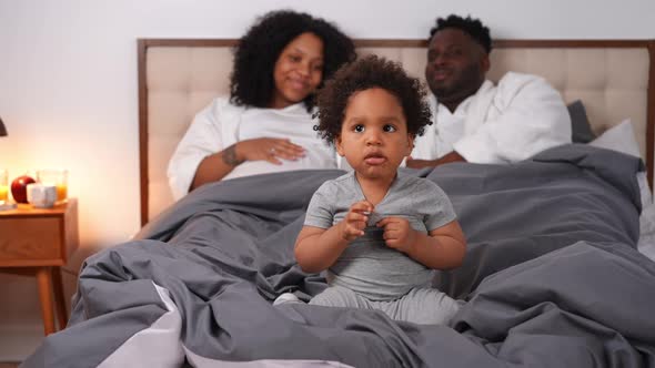 Portrait of Cheerful Cute Little African American Boy Sitting on Bed with Parents Talking at