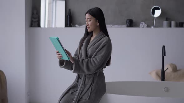 Young Gorgeous Asian Woman in Bathrobe Scrolling Tablet Screen Surfing Internet in Bathroom Indoors