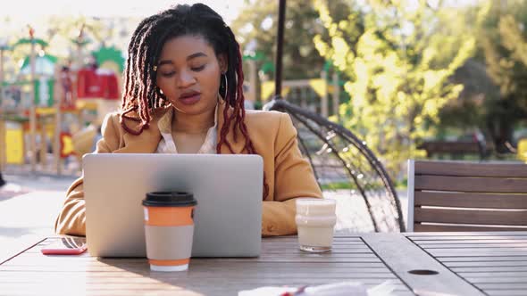 Young African American Woman Sitting in Outdoor Cafe and Using Laptop