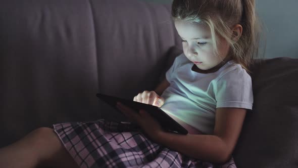 Little preschool girl use digital tablet technology device lying at sofa in dark room