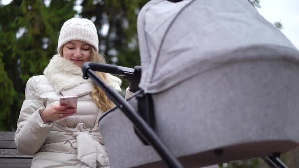 Happy Mother with Smartphone Using App. She Sitting on the Bench , Baby Sleeping in Pram