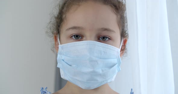 A Child at Home in a Protective Medical Mask with a Sad Face and Eyes Looks at the Camera