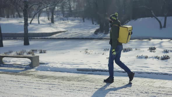 Side View of Courier in Medical Mask with Thermo Bag Walking Along Snowy City Street