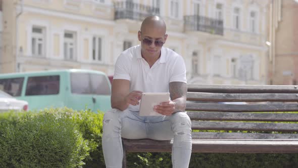 Handsome Bald Middle Eastern Man Wearing Sunglasses and White T-shirt Typing on Tablet