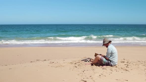 A man in a blue shirt and a hat reading a bible on the beach by a small fire.