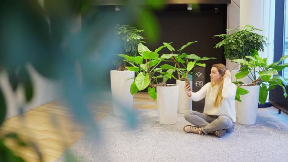 a Beautiful Woman with Long Blond Hair Sits on the Floor Cuts Green Plants and Talks on a Smartphone