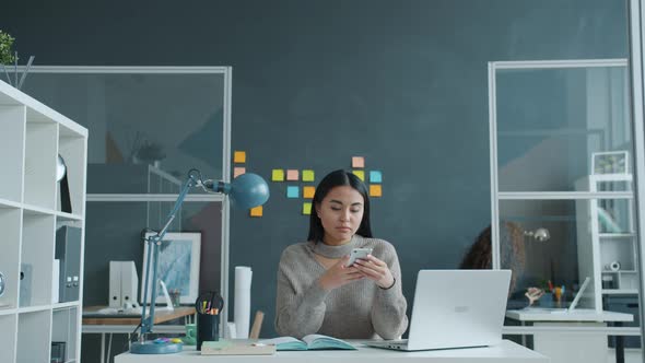 Happy Asian Girl Using Smartphone Smiling Texting at Work While Woman Is Working in Background