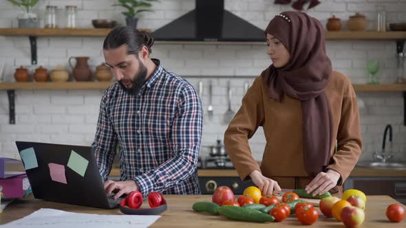 Young Beautiful Wife in Hijab Cutting Cucumber in Kitchen As Busy Husband Surfing Internet on Laptop