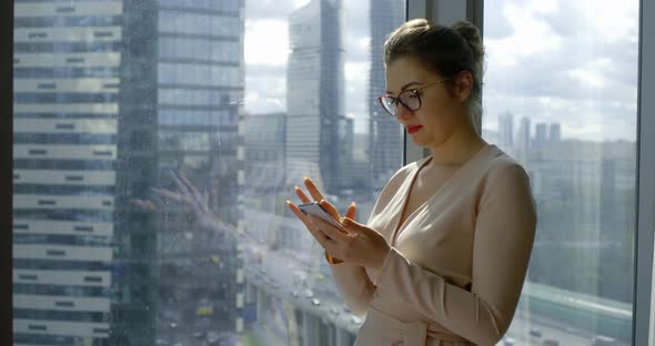 Young Brunette in Glasses Holds Modern Smartphone and Types