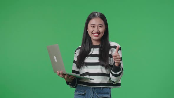 A Smiling Asian Woman Showing Thumbs Up Gesture While Typing On Computer In The Green Screen Studio