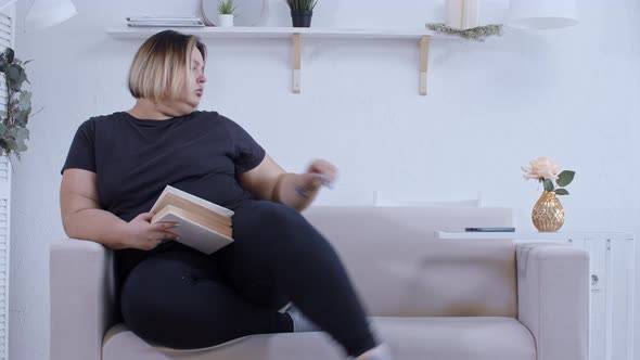 A Fat Woman Lying on a Couch Reading a Book and Checking Her Phone at Home