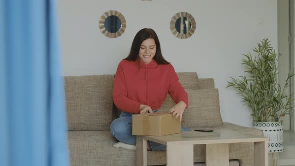 Satisfied brunette woman in red hoodie shaking the box and opening the package at home